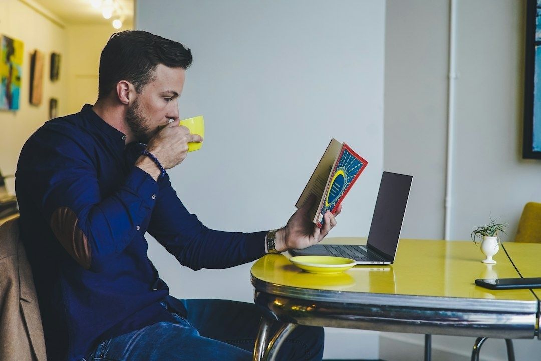 Mann leser en bok og drikker te ved et bord med en bærbar PC